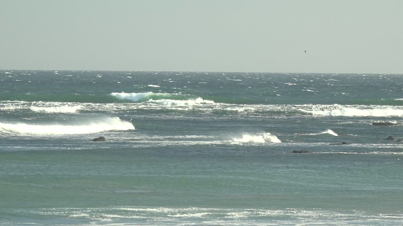 At 0:02 in La Bocana, Baja California Sur, wind-roughened water fills the frame as bright white surf rolls over dark rocky areas under flat gray light, with no visible people.