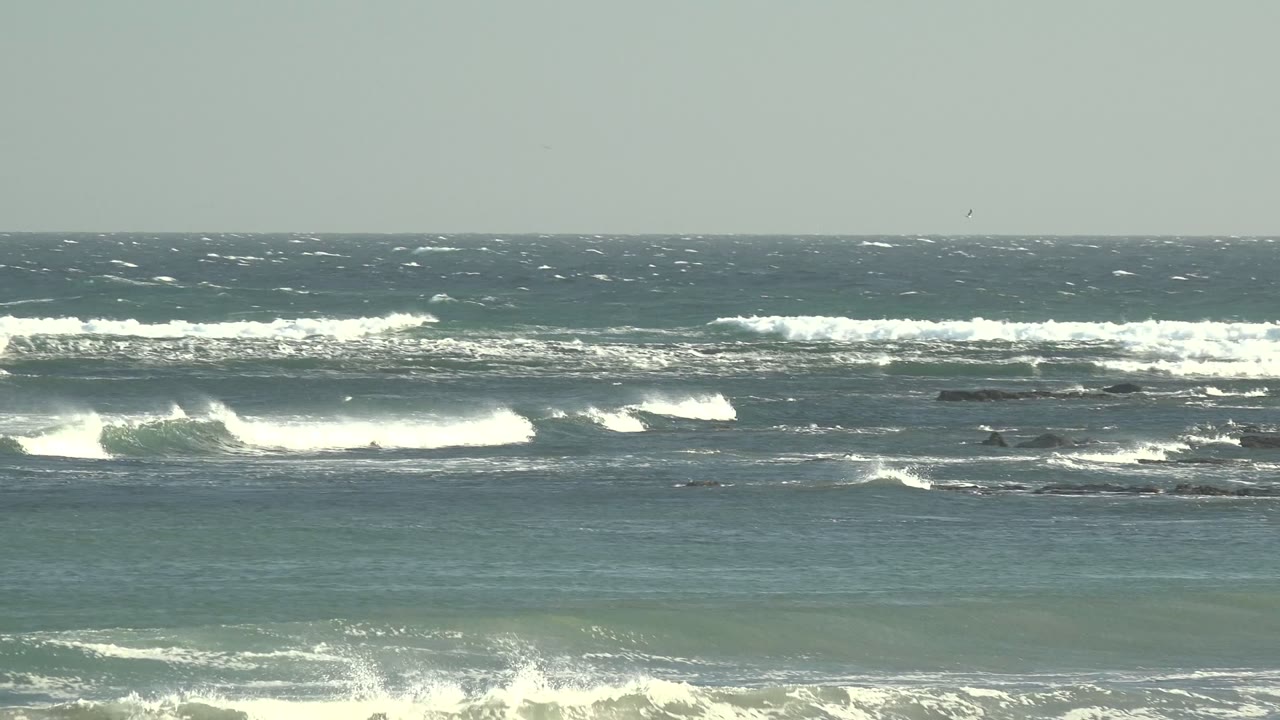 Wide La Bocana ocean view with dark rocks across the surf line, white waves breaking in bands, blue-green water in the foreground, and no visible people.