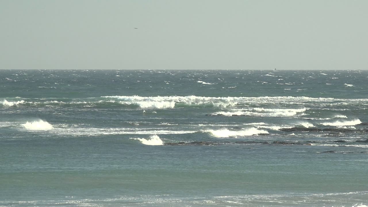 La Bocana, Baja California Sur ocean view with whitecaps breaking across dark rocks, pale gray sky above, and no visible people.