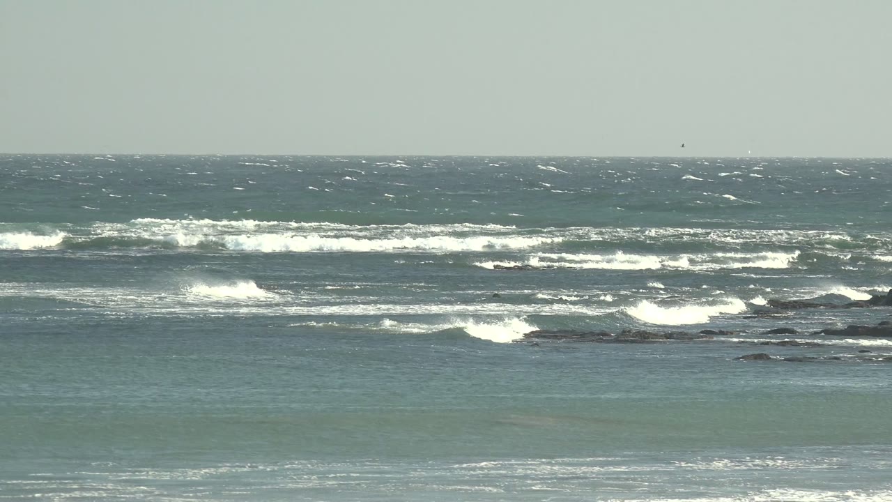 La Bocana coastline with layered white surf lines, blue-green shallows near the bottom, dark rocks in the midground, and no visible people.