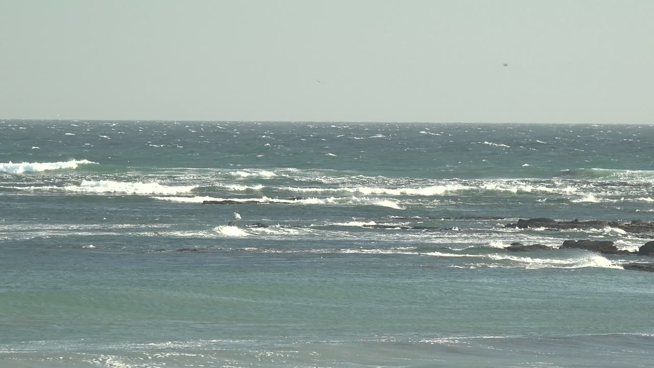 Wide view of La Bocana, Baja California Sur ocean surf with whitecaps breaking over scattered dark rocks beneath a pale gray sky, no visible people.