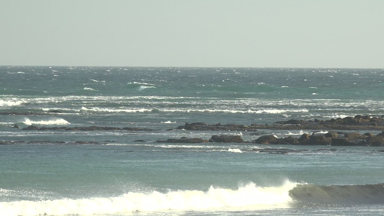 A broad white surf line breaks across the La Bocana shoreline with low rocks behind it and textured blue green water beyond, with no visible people.
