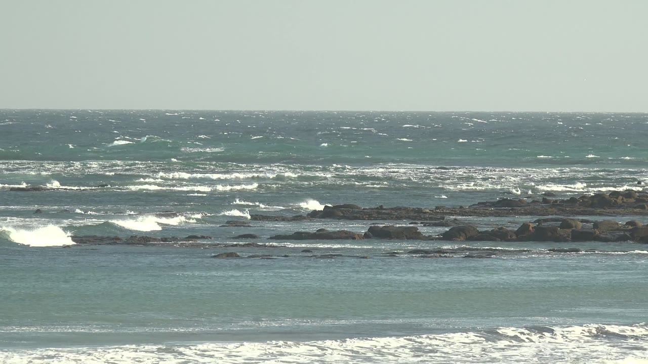 Wide view of choppy blue-green ocean at La Bocana, Baja California Sur, with whitecaps, low rocks, foreground foam, and no visible people.