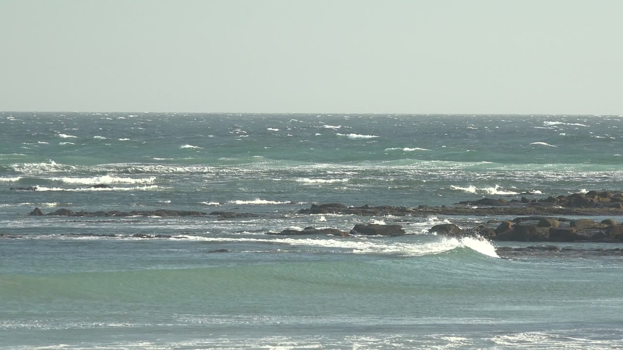 Wide view of waves breaking across rocks at La Bocana, Baja California Sur, with blue-green water, pale sky, and no visible people.