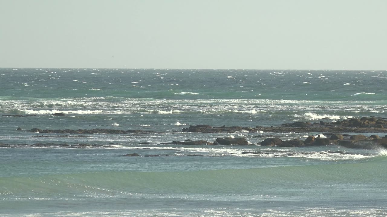 A wide coastal frame shows white surf lines crossing low rocks and darker offshore water under a clear pale sky in La Bocana, with no visible people.