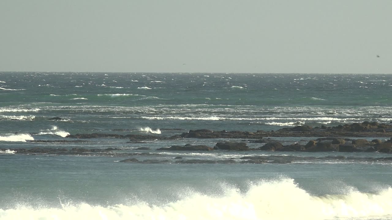 Wide view of waves breaking across dark rocky formations at La Bocana, Baja California Sur, Mexico, with pale sky, choppy water, and no visible people.