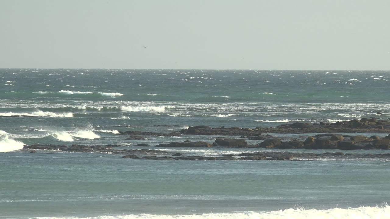Choppy open water and whitecaps stretch behind the rocky La Bocana shoreline, while foamy surf rolls through the foreground and no visible people appear.