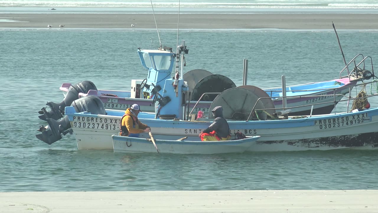 At La Bocana, Baja California Sur, a person in an orange shirt works an oar from a small skiff beside anchored blue and white fishing boats, with another person seated nearby.