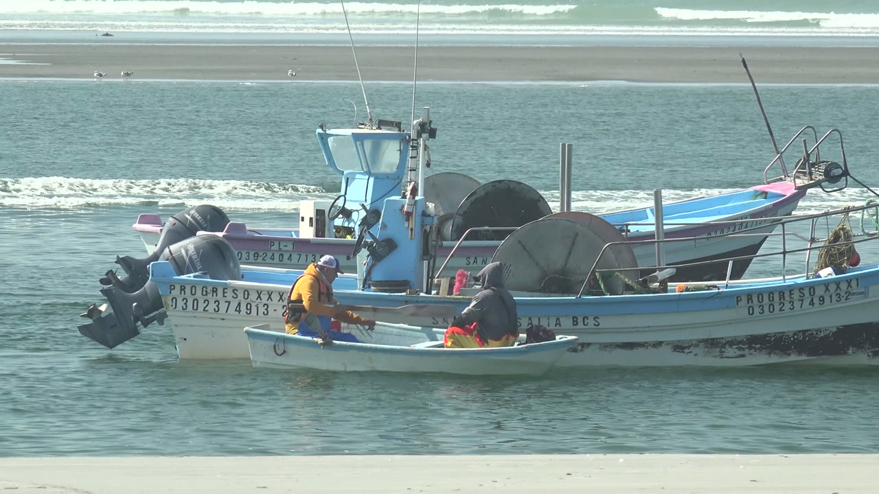 Small fishing boats float beside larger blue and white boats at La Bocana, Baja California Sur, with fishermen aboard and surf breaking beyond the sandbar.