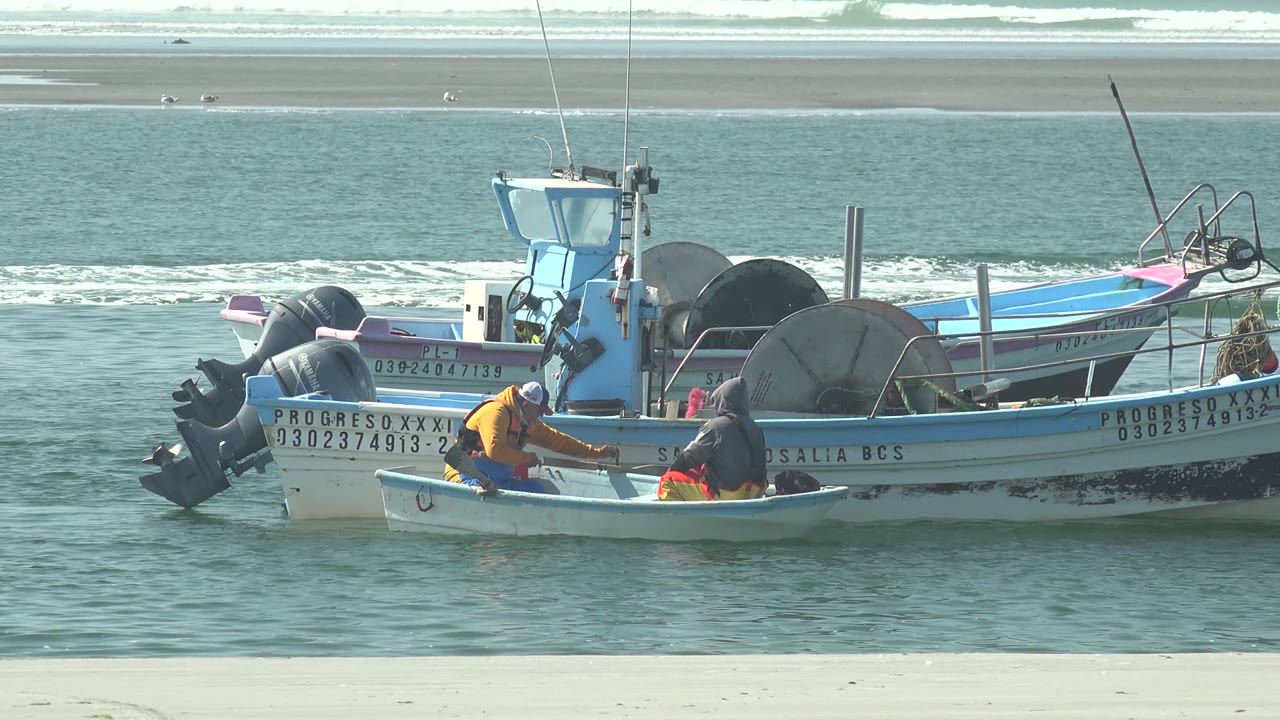 At La Bocana, fishermen in a small white boat work next to larger blue-trimmed fishing boats, with pale beach and low ocean waves behind them.