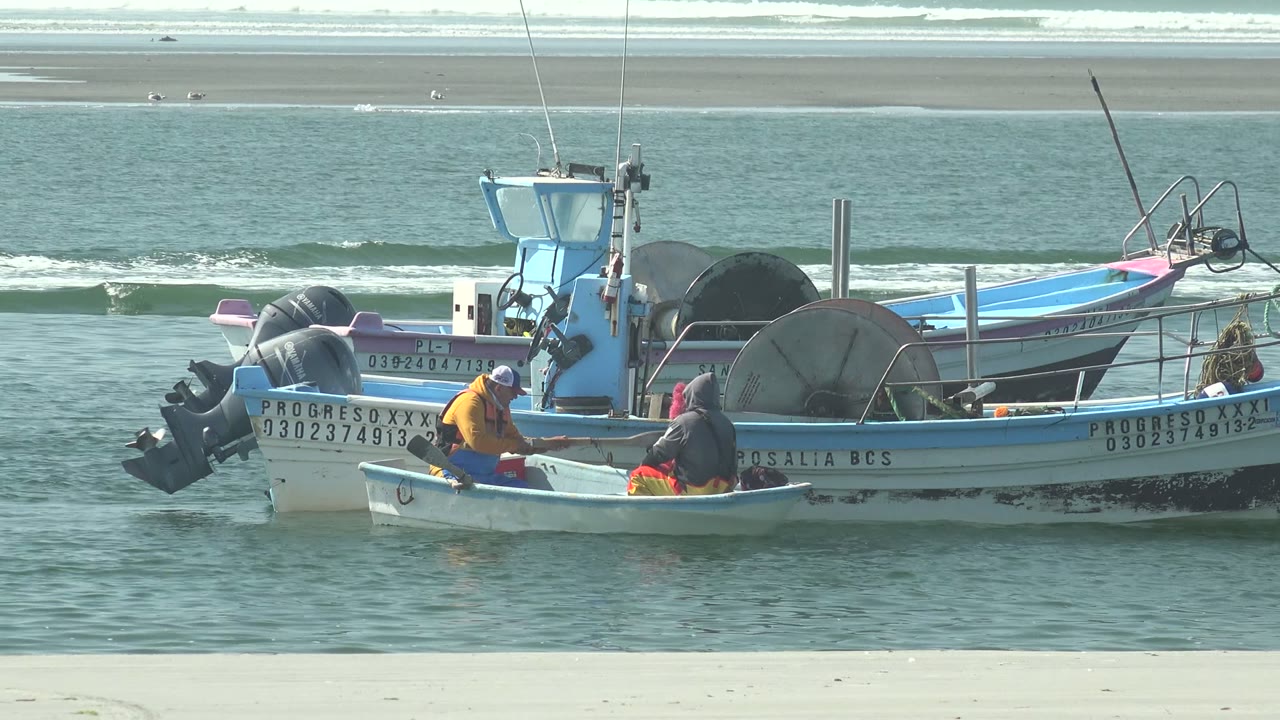 Two fishermen remain low in a small boat at La Bocana while a row of larger fishing boats, outboard motors, and calm shoreline water fill the frame.