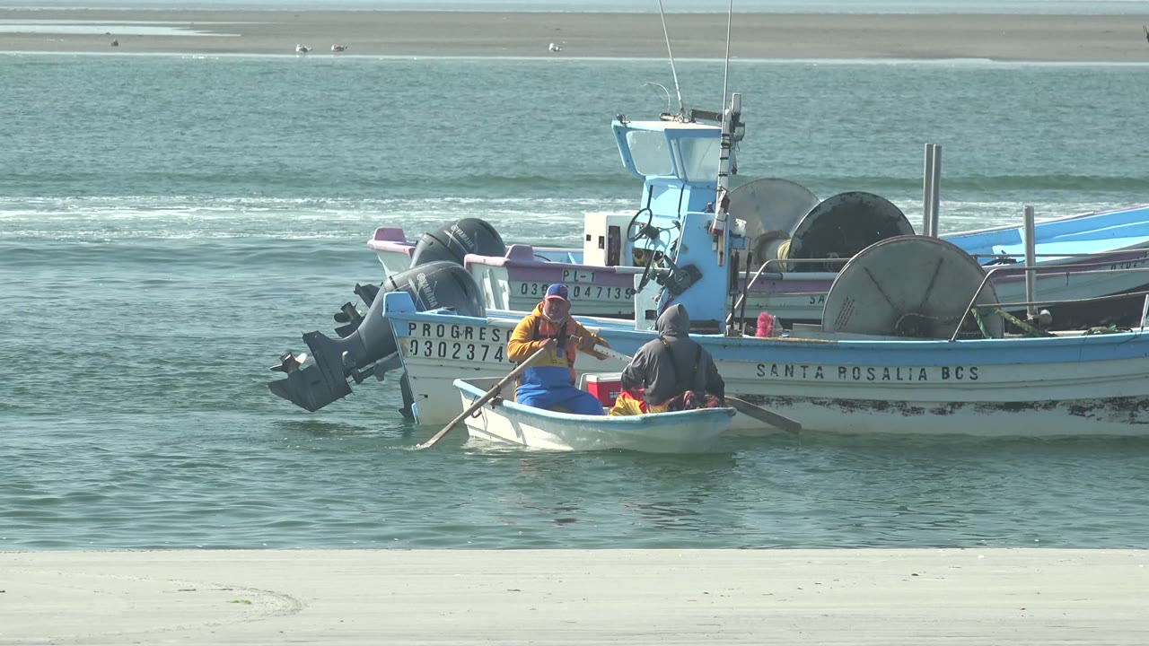 Two people in a small rowboat sit close to a larger blue-and-white fishing boat in La Bocana, Baja California Sur, with sandy shore and low waves behind them.