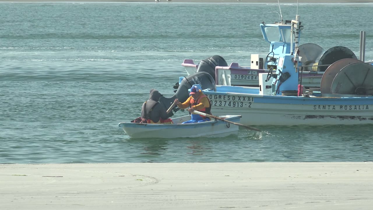Two people remain in the small rowboat near the stern and outboard motors of a fishing vessel in La Bocana, Baja California Sur, with the oars angled across the water.