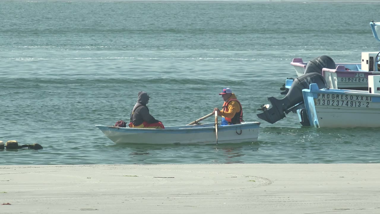 At 0:00 in La Bocana, Baja California Sur, two people sit in a small white boat near sandy shore while a larger motorboat with visible lettering and numbers sits behind them.
