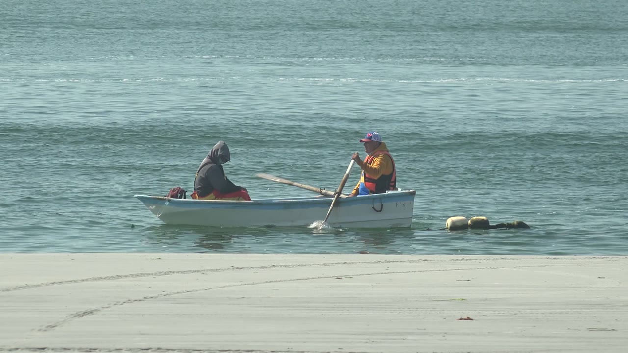 Two people in a small white rowboat sit near the sandy shore at La Bocana, Baja California Sur, with one person facing the water and another holding an oar.