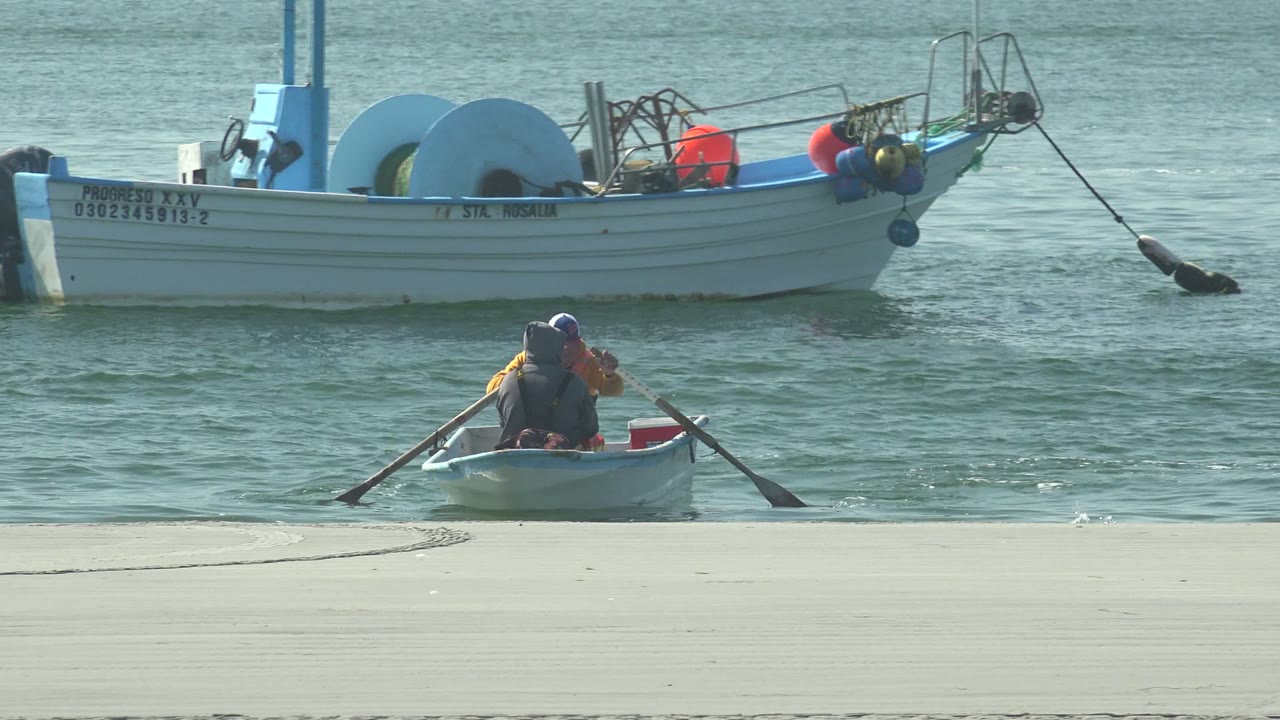 At 0:00 in La Bocana, two people row a small white dinghy near the sandy edge while a larger white fishing boat sits behind them on calm water.