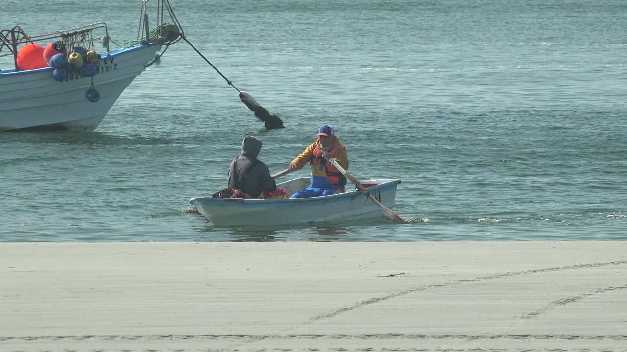 At 0:00 in La Bocana, Baja California Sur, two people row a small white boat near the beach while a larger boat sits anchored in the background water.