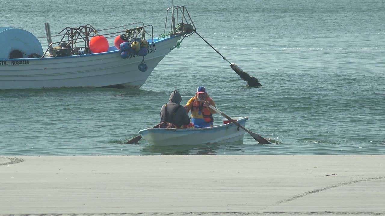 At 0:02 in La Bocana, Baja California Sur, the two-person rowboat is farther right, with oars extended and the larger fishing boat broadside behind it.