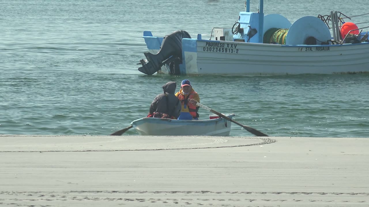 At 0:00 in La Bocana, Baja California Sur, two people sit in a small rowboat just off the sand while a larger boat floats behind them in green-blue water.