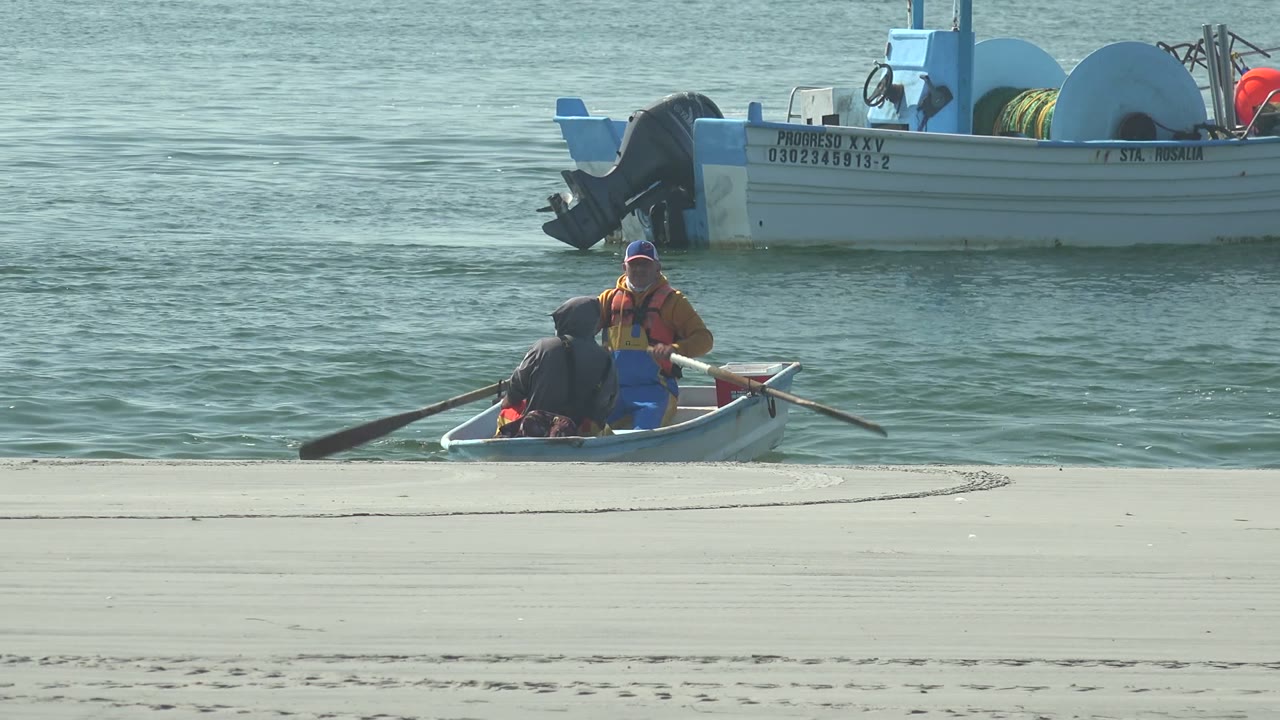 Rowboat with two people near pale sand and a larger boat in the water behind.