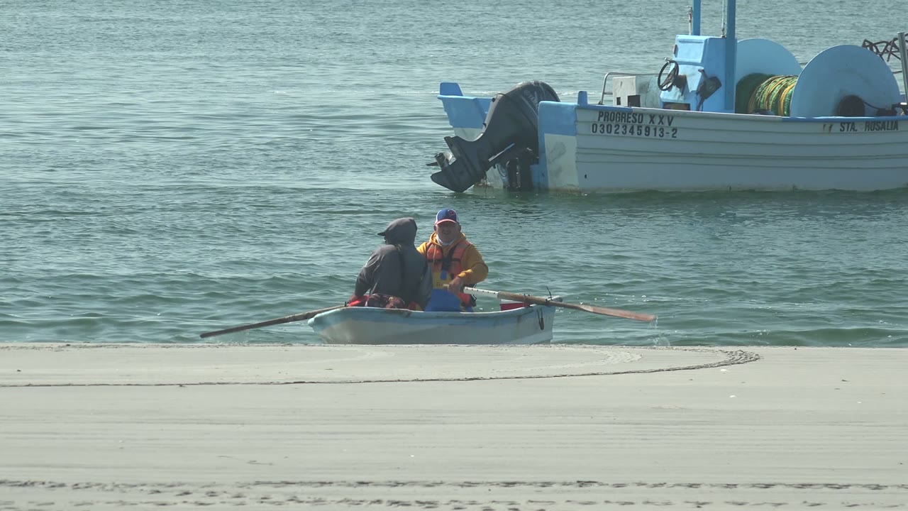 At 0:02 in La Bocana, Baja California Sur, the rowboat has shifted right in the frame, with both oars extended over the water and the larger boat cropped in the upper background.