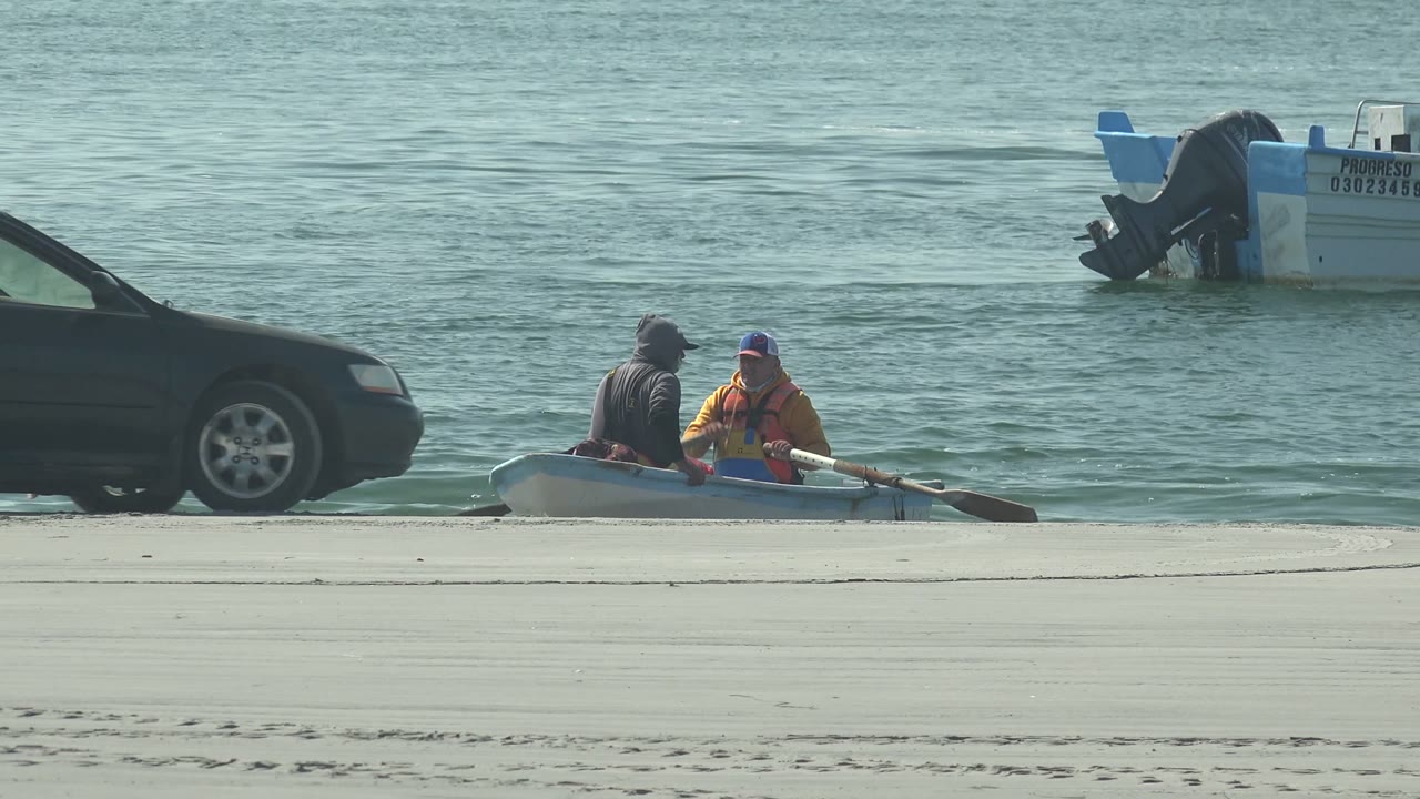 At La Bocana in Baja California Sur, a dark car pulls a small inflatable boat with two visible people along the edge of the water, with a motorboat offshore behind them.