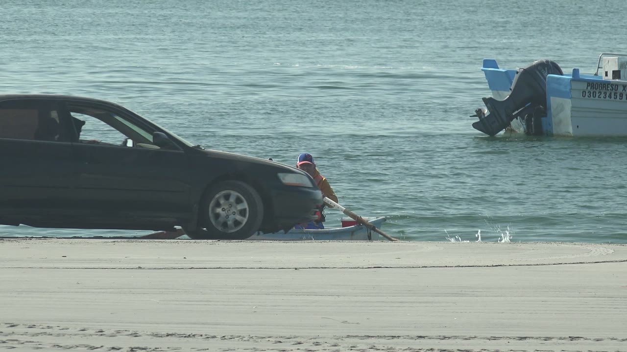 The sedan continues rightward along the La Bocana beach as one person in bright outerwear remains visible beside the small boat at the edge of the water.