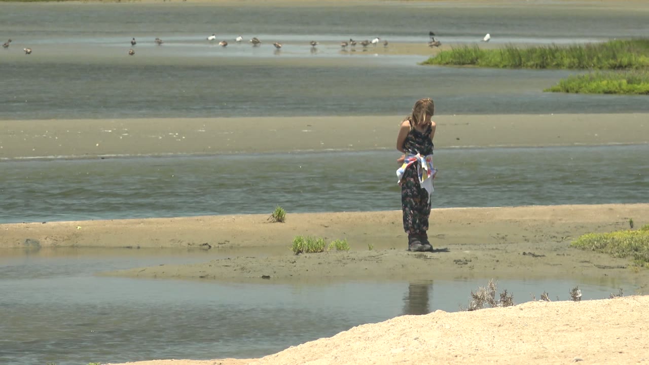 At Guerrero Negro, Baja California Sur, the person faces away on the wet shore, with the white towel visible at the waist and a band of shallow water, sandbar, and birds in the background.