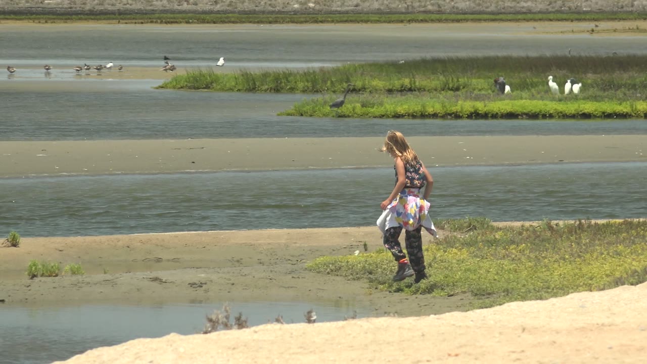 Girl walking across the muddy shoreline with birds in the wetland behind her.