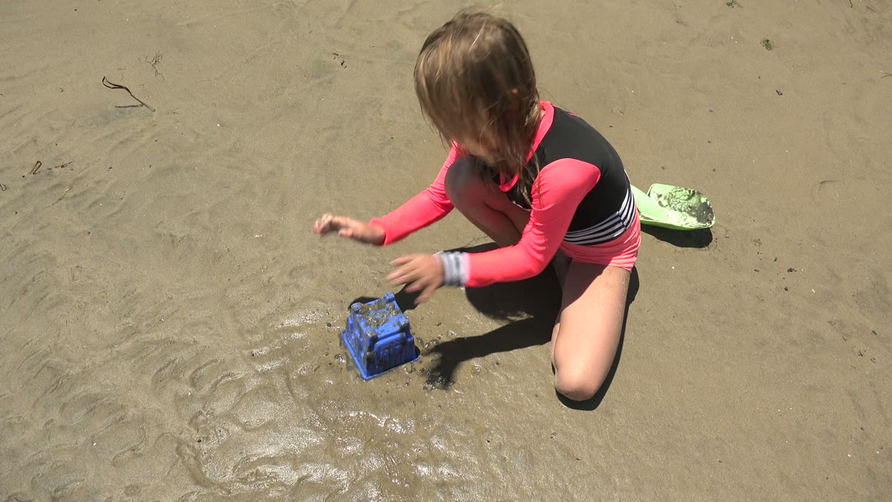 At 0:00 in San Carlos, Mexico, a child in a pink and black long-sleeve top kneels over wet sand with one hand lifted near a blue sand toy.