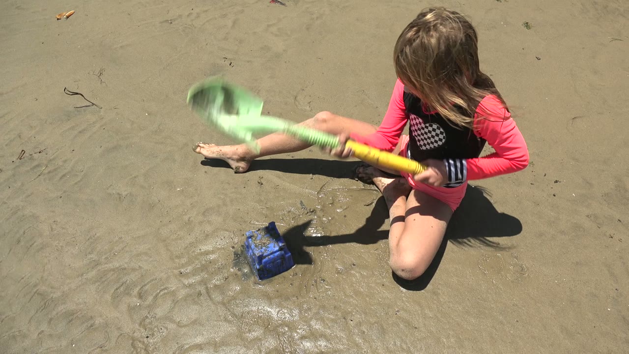 A young girl in a pink and black swimsuit sits on wet sand in San Carlos, Mexico, holding a green shovel beside a small blue bucket.