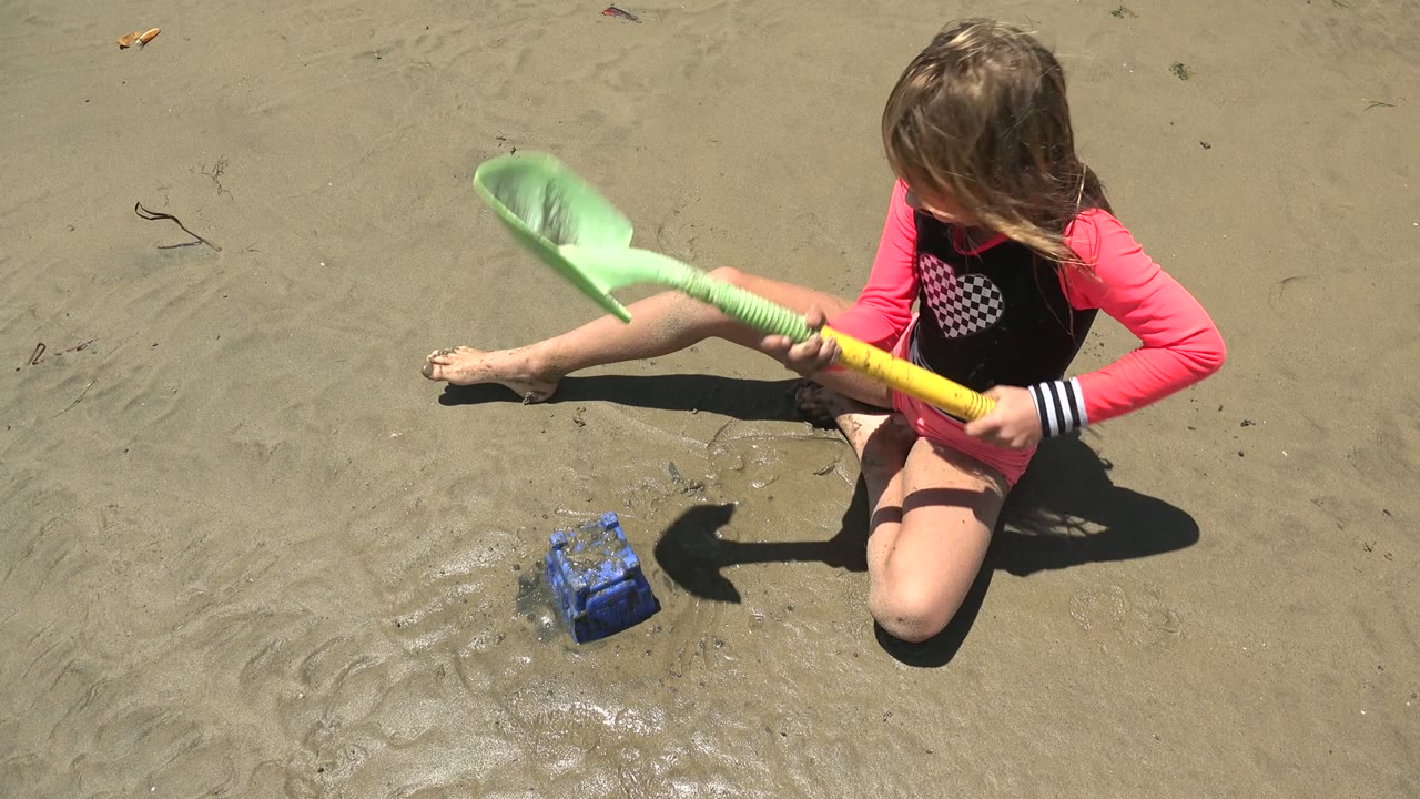 Girl swings a green shovel across wet beach sand beside a blue bucket.