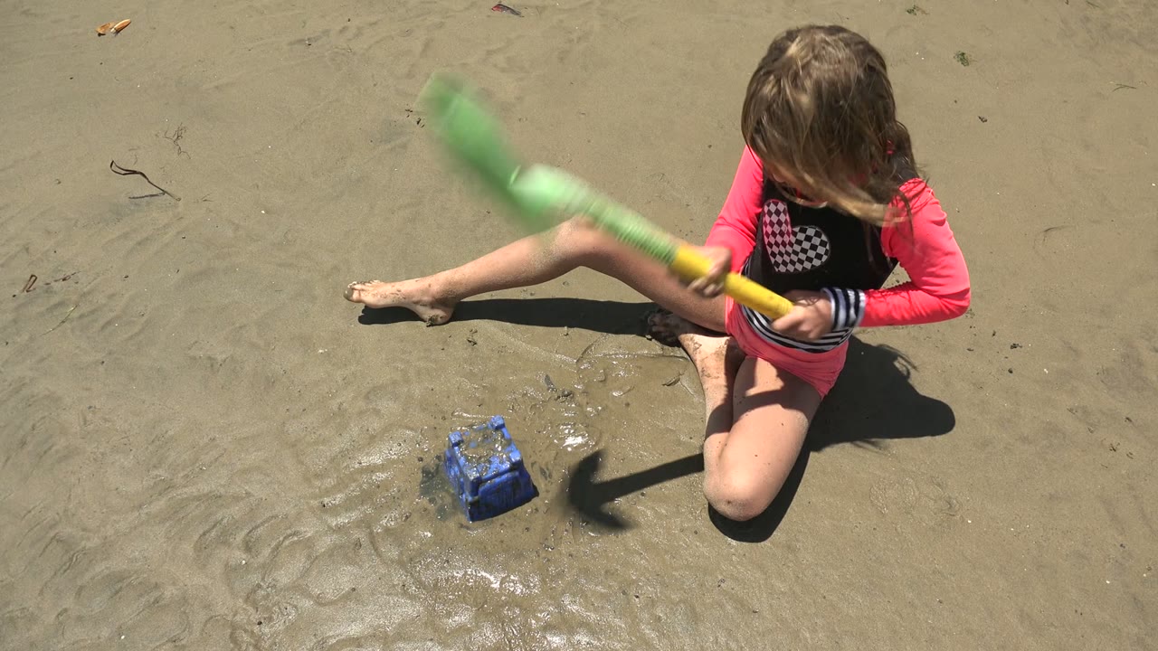The girl lifts the green shovel with visible motion blur while the blue bucket remains on the wet sand in San Carlos, Mexico.