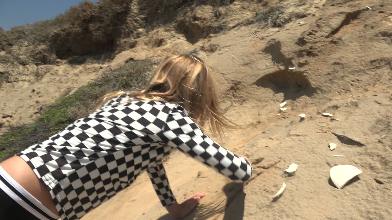 Girl crouching beside white shell pieces on a sandy slope.