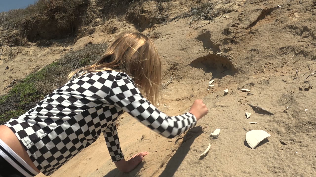 The girl extends her arm toward several white shell-like fragments on the sandy San Quintin hillside while her hair falls forward over her face.