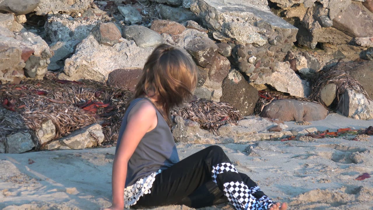 A child sits low on the sandy beach in San Carlos, Mexico, with rocks and dried seaweed behind her in warm late-afternoon light.