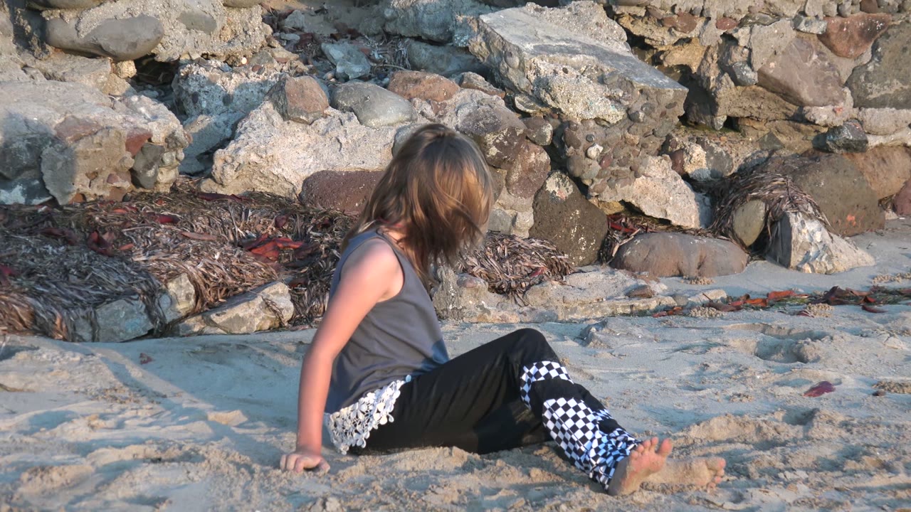 Child seated on sand with rocky shore behind her.