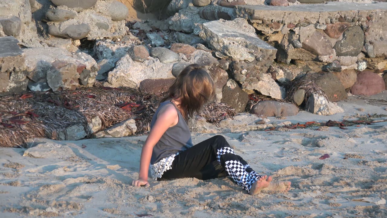 A seated child on the San Carlos shoreline faces toward the rocks, with bare feet on rippled sand and seaweed gathered along the stone edge.