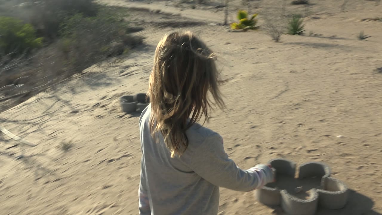 A girl seen from behind walks across sandy ground in San Carlos, Mexico, near grouped tire structures, low cactus, scrub, and bright afternoon light.