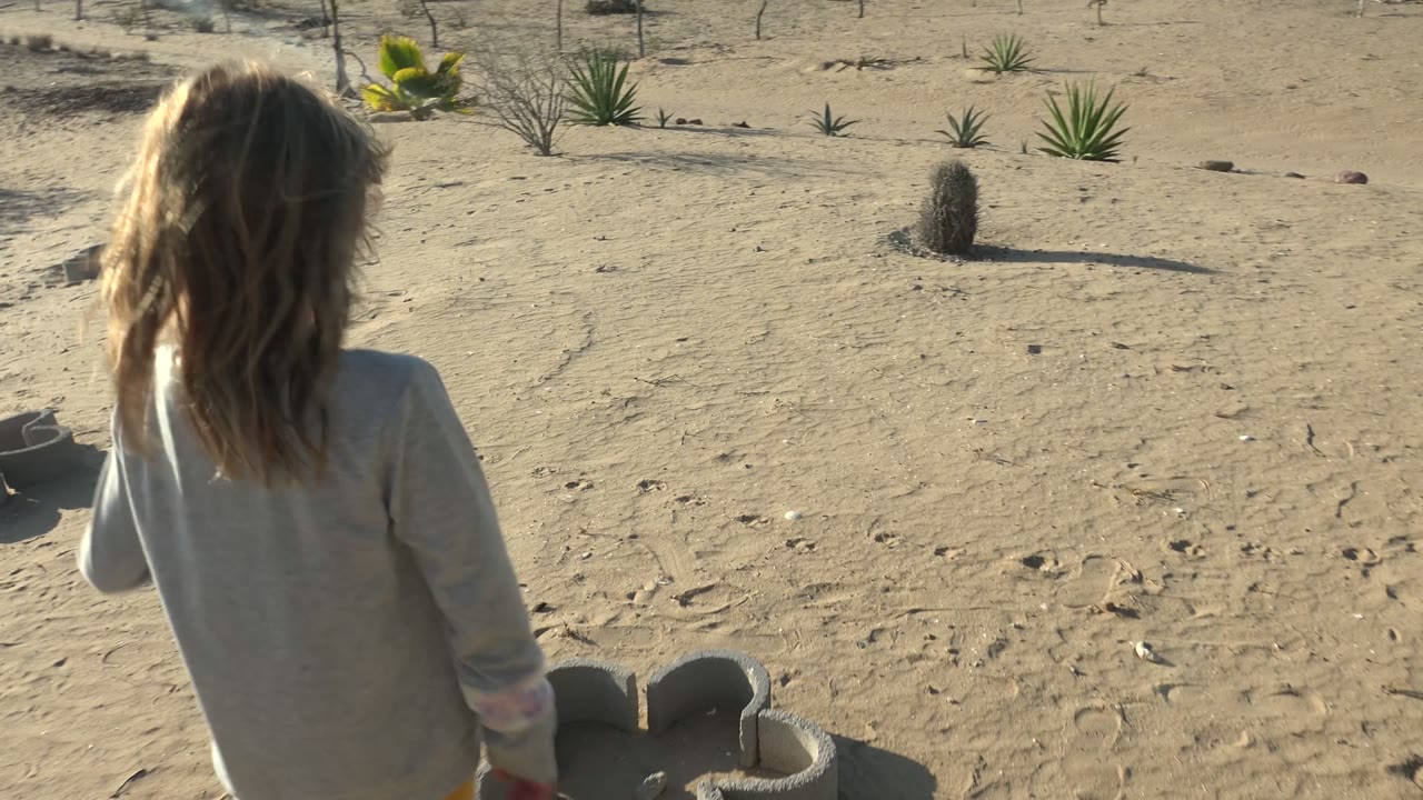 Girl walking away on sand with small cactus and tire structures nearby.