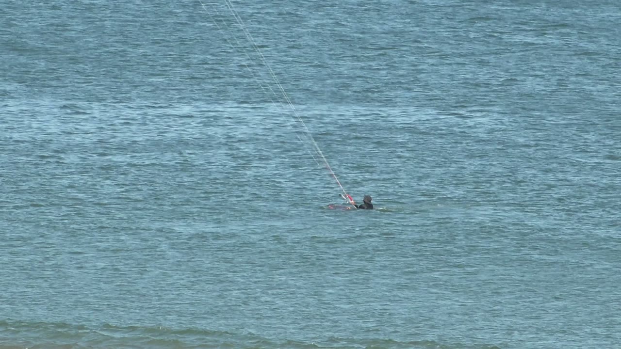A distant kite surfer in San Juanico, Mexico glides near the upper right of the frame as a low wave line crosses the blue water behind the rider.