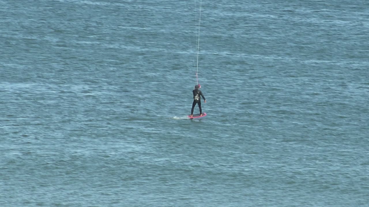 A distant kite surfer in a dark wetsuit rides a red board on rippled blue water in San Juanico, Baja California Sur, with kite lines rising out of frame.