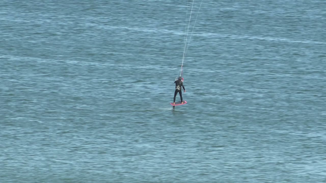 A small kite surfer on a red board appears against a broad field of textured blue water in San Juanico, Baja California Sur, with the kite itself outside the frame.