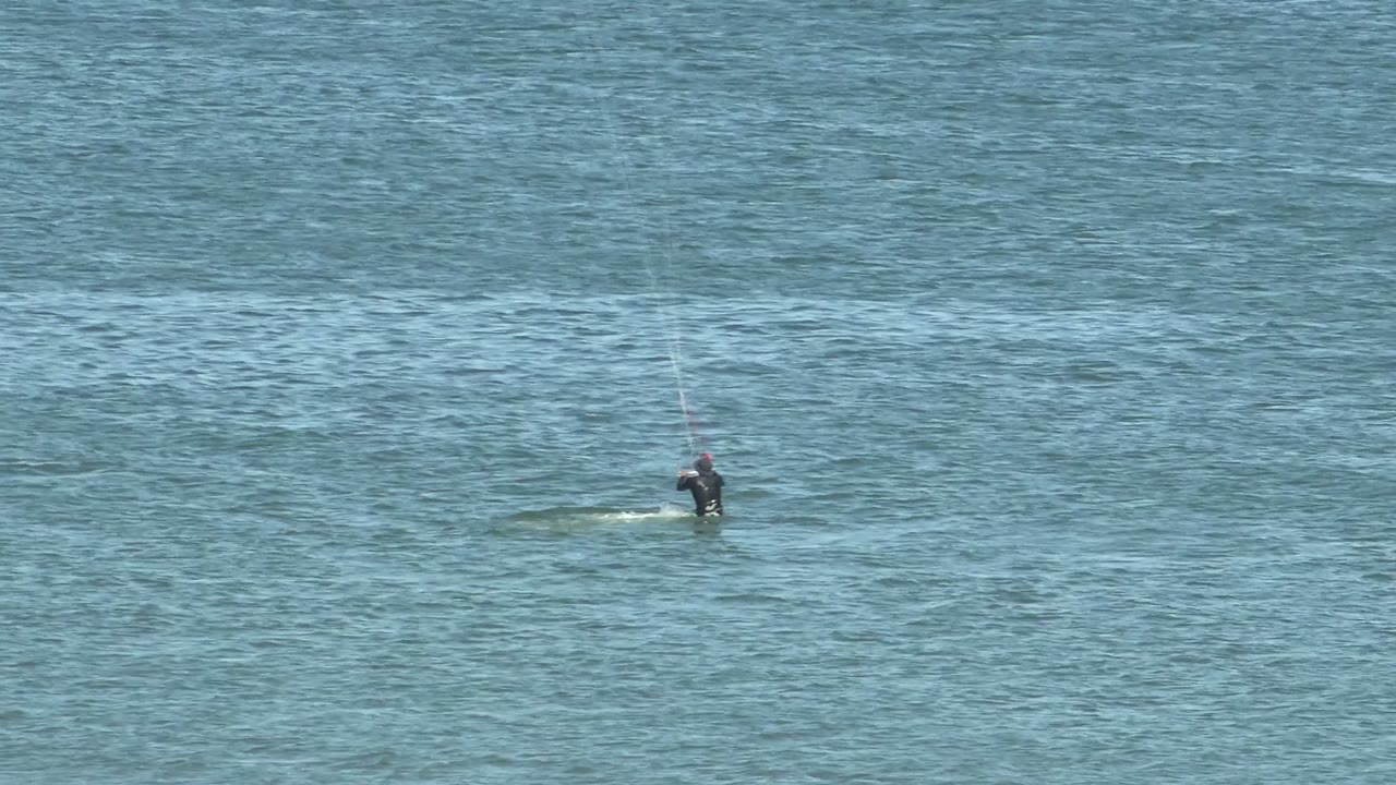 A distant kite surfer rides low across blue ocean water in San Juanico, Baja California Sur, with kite lines faintly visible above the rider.