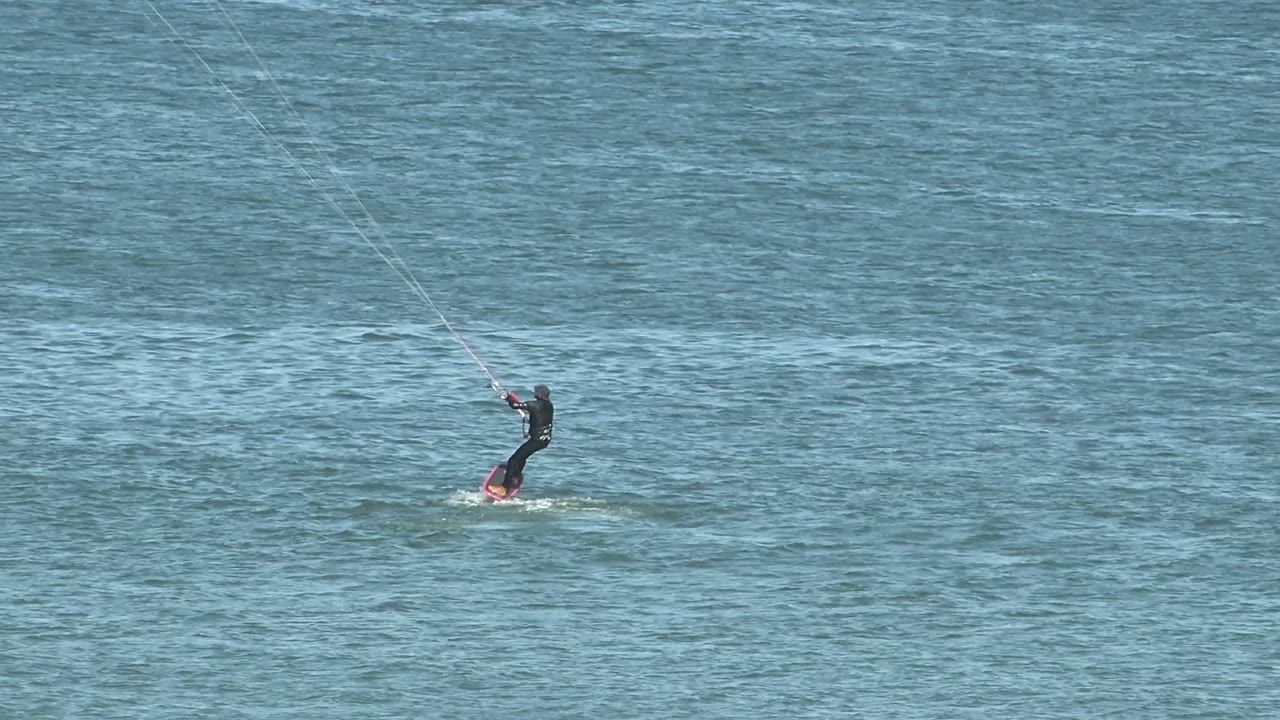 A distant kite surfer glides over the ocean in San Juanico, Baja California Sur, with a pink-toned board visible beneath the rider.