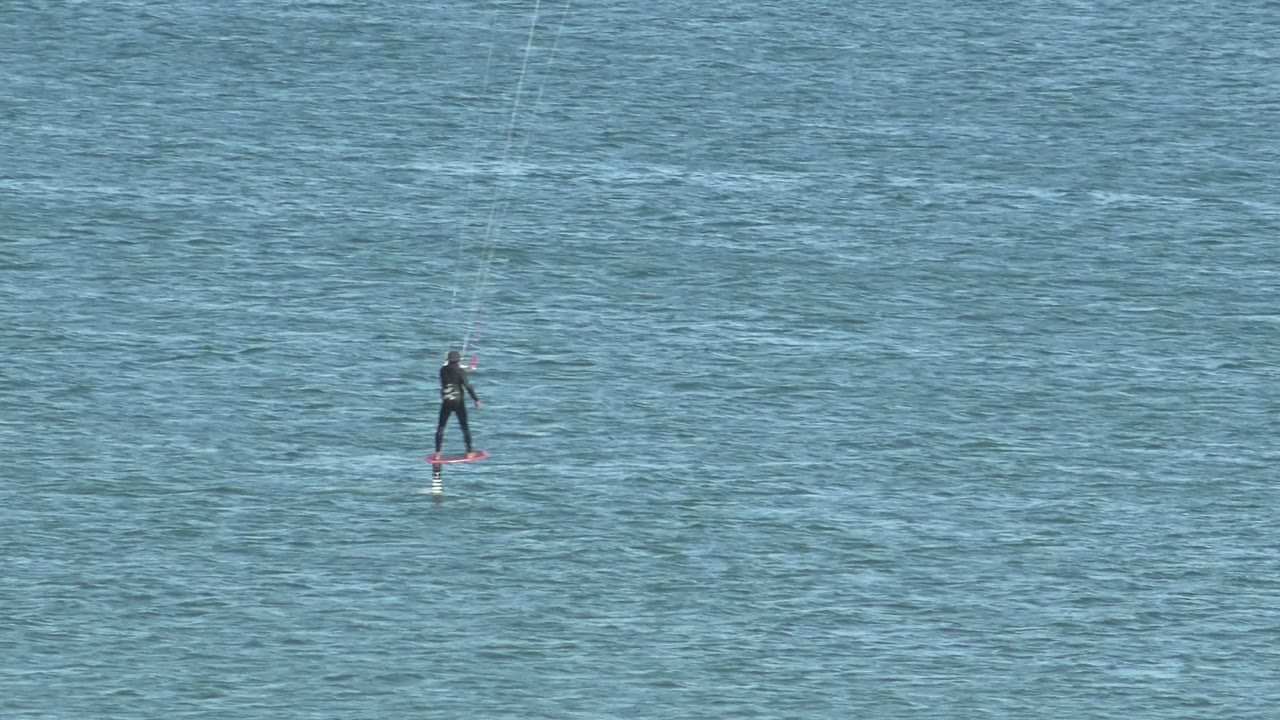 At 0:00, a distant kiteboarder in San Juanico rides a red board across blue water with thin kite lines extending upward out of frame.