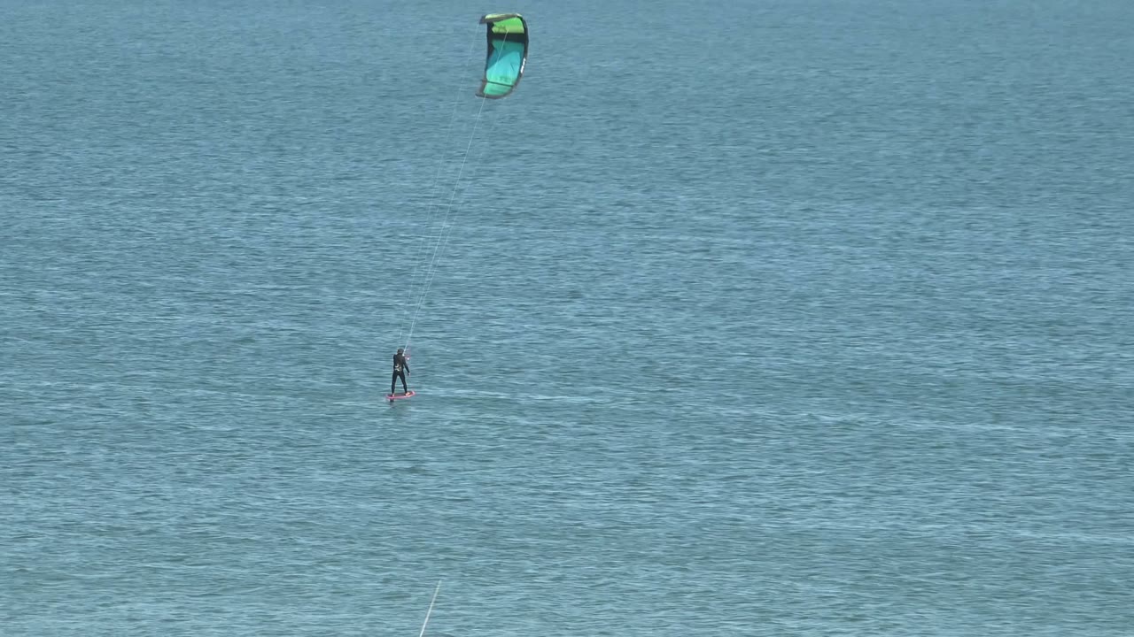 A distant kite surfer rides on blue ocean water in San Juanico, Baja California Sur, with a green and blue kite high above the rider and a faint horizon line in the background.
