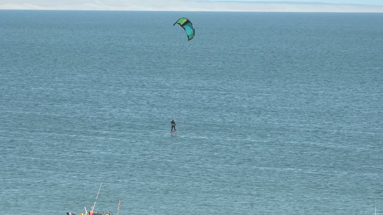 A small rider moves across the water in San Juanico while the green kite sits near the upper right of the wide ocean frame, with no close foreground detail visible.