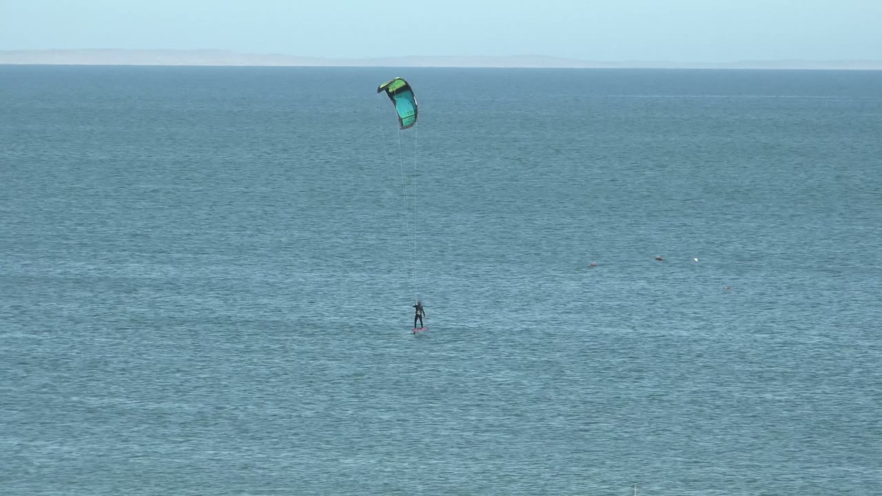 Wide San Juanico ocean frame with a distant kite surfer below a green and blue kite, blue water filling the view, and a pale horizon line in the background.