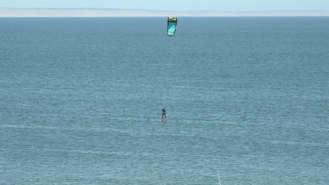 A distant kite surfer rides on open water in San Juanico, Baja California Sur, with a green and blue kite high above and a pale horizon near the top of frame.